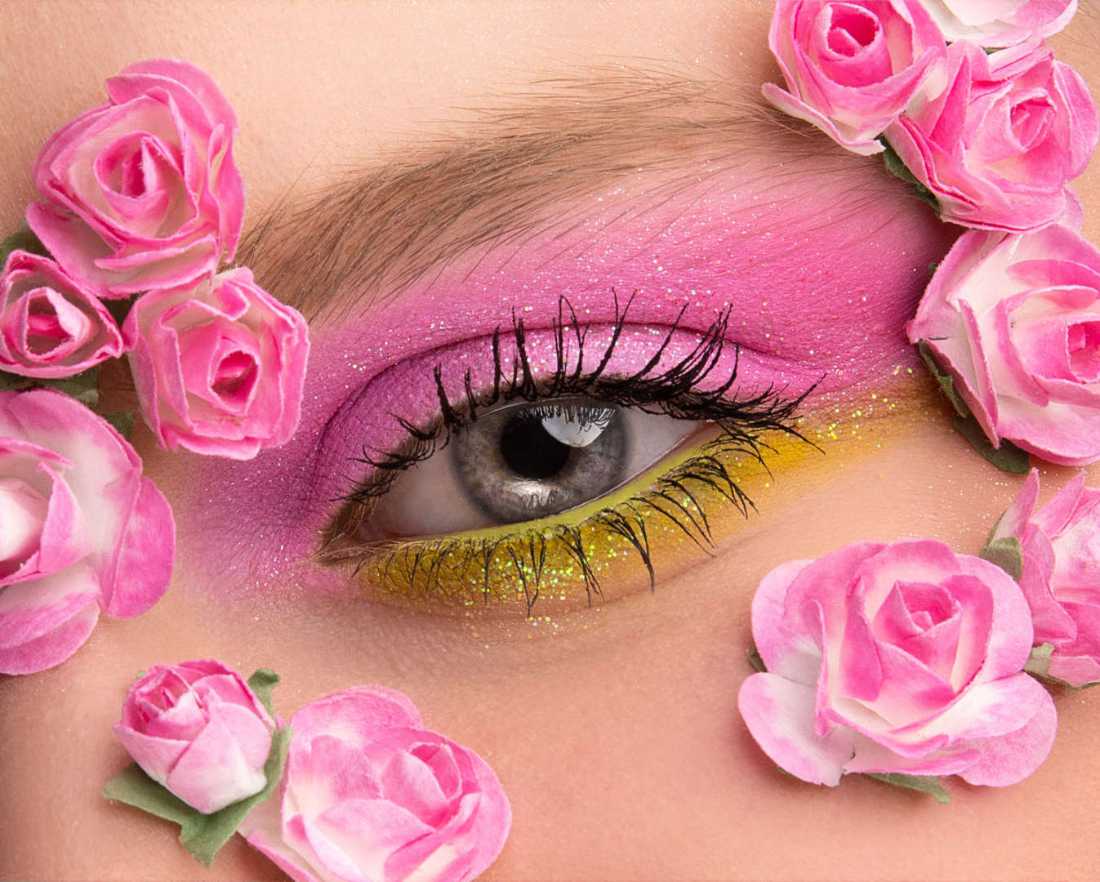 Fotografía close-up de un ojo color azul con maquillaje rosa y verde y flores a su alrededor como adorno.
