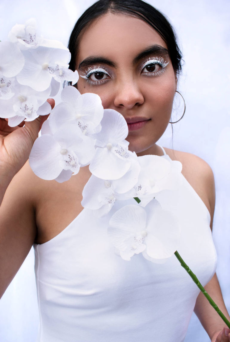 Fotografía en plano medio de una joven con maquillaje y atuendo blanco sosteniendo un racimo de orquídeas.