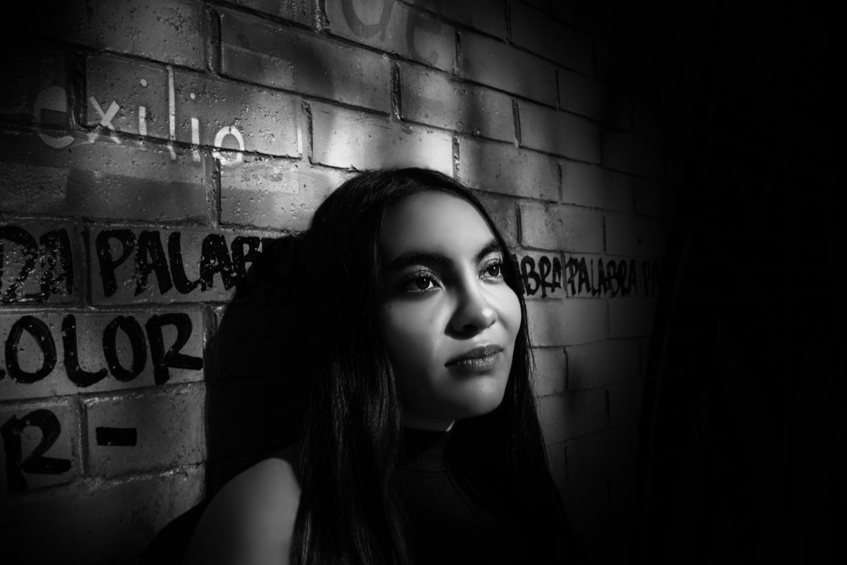 Fotografía primer plano en blanco y negro de una joven recargada en una pared con grafitis, destacando uno que dice «exilio».
